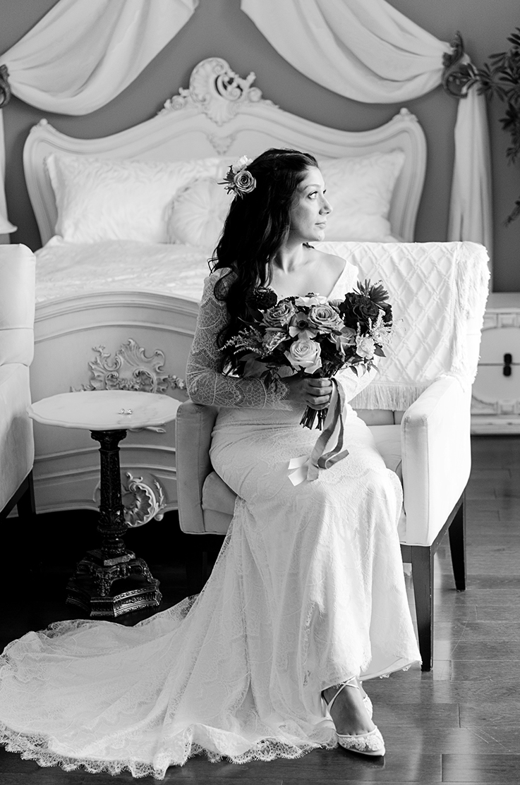 A bride in a long sleeve lace wedding dress sits on a chair holding a bouquet, while looking off to the side in a bedroom.