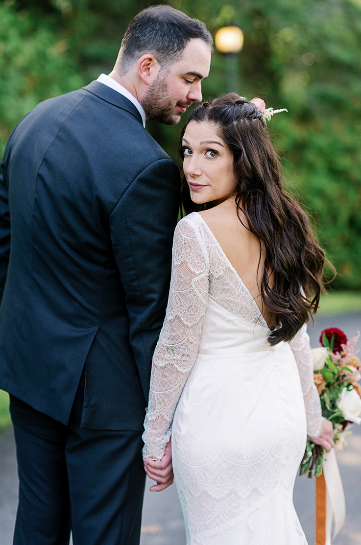 A bride in a long sleeve lace wedding dress and a groom in a suit stand and look back towards the camera, while outside.
