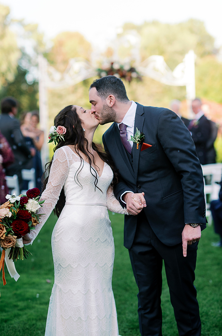 A bride in a long sleeve lace wedding dress and a groom in a suit kiss outside after their wedding ceremony.