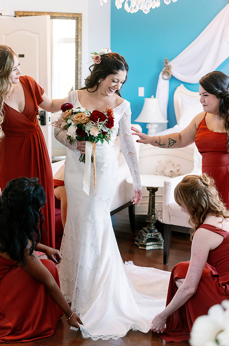 A bride in a long sleeve lace wedding dress smiles and holds a bouquet while her bridesmaids help her with her dress in a hotel room.