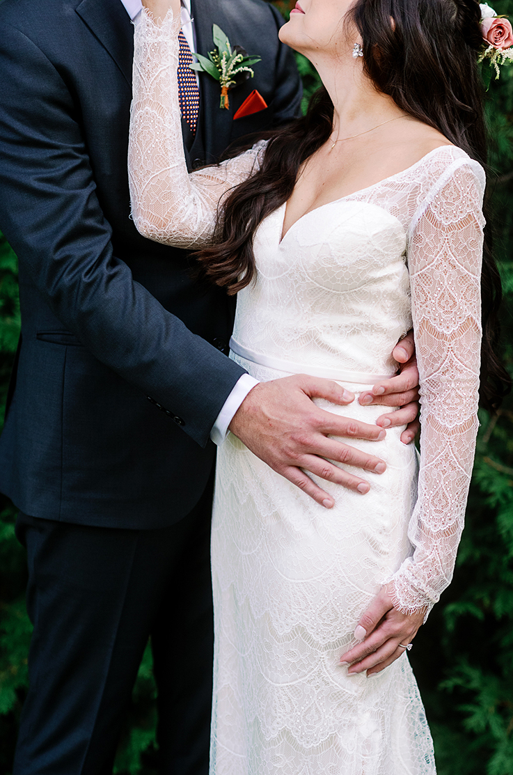 A cropped image of a groom in a suit holding the waist of a bride in a long sleeve lace wedding dress.