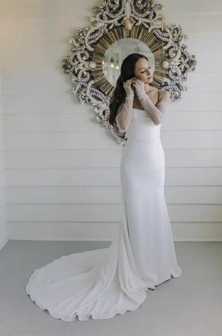 Bride in a scoop neck crepe gown with sheer sleeves, puts on earrings by an ornate mirror on a white wall. 