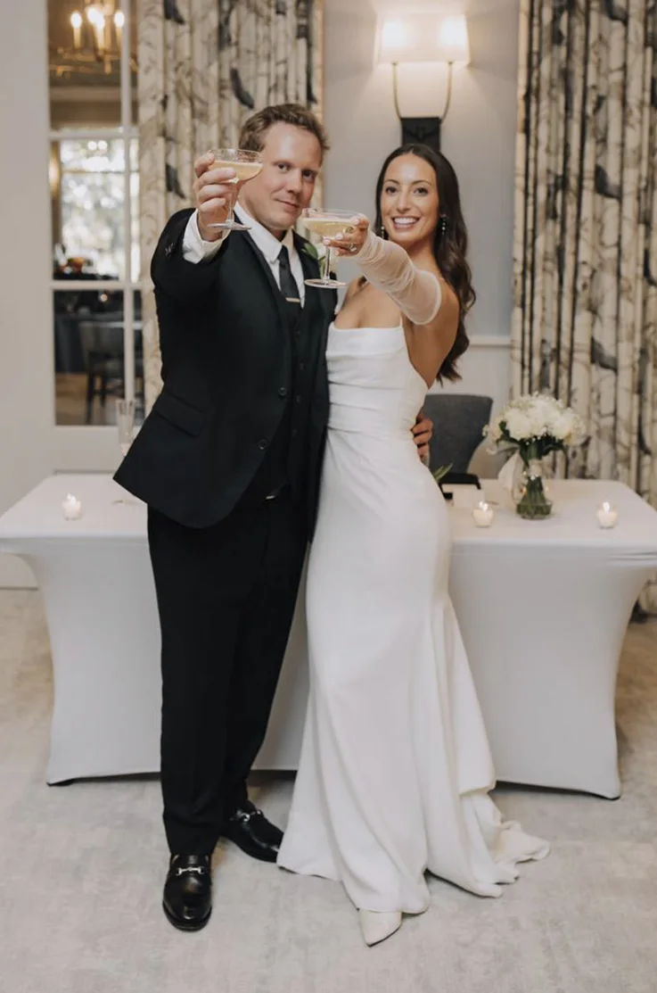 Bride in a sleek crepe wedding dress with a pleated bodice, smiles holding up drinks with the groom. 