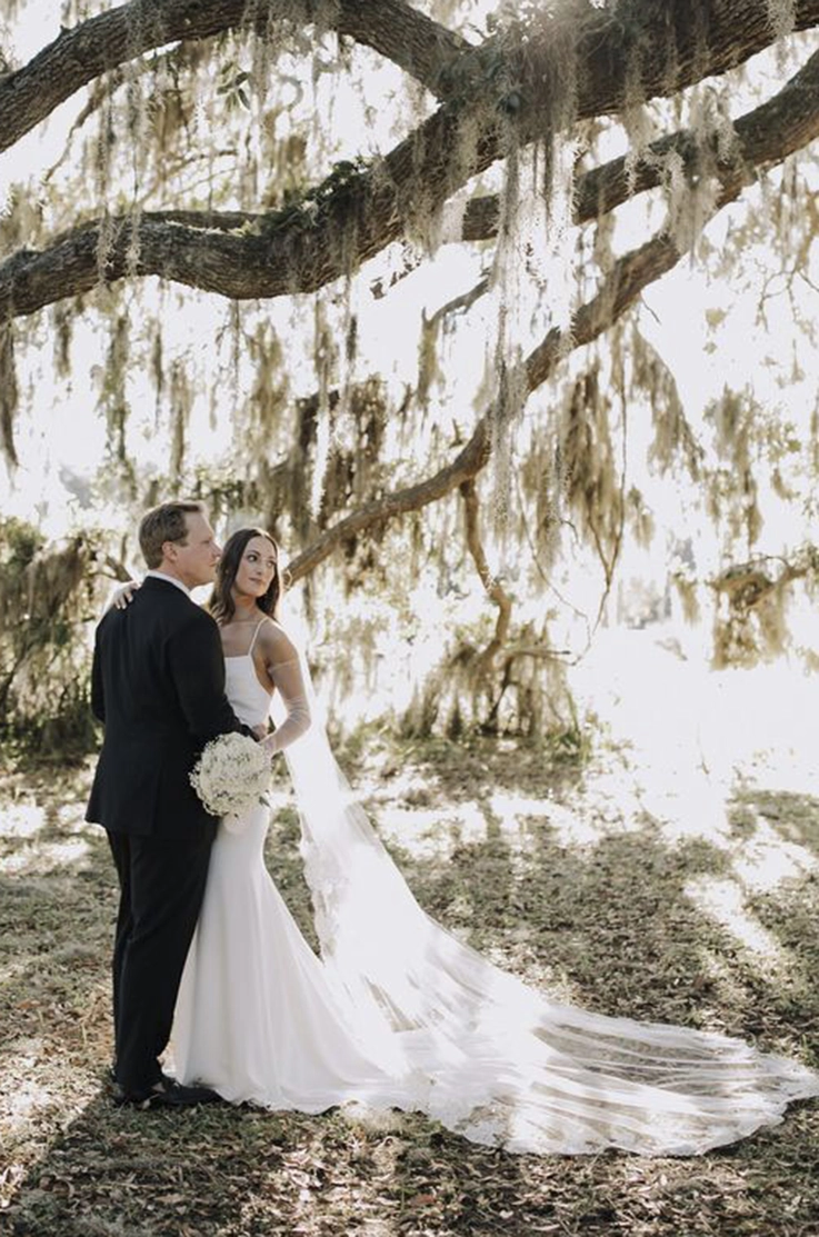 Groom embraces bride in a spaghetti strap crepe gown under trees with hanging leaves. 