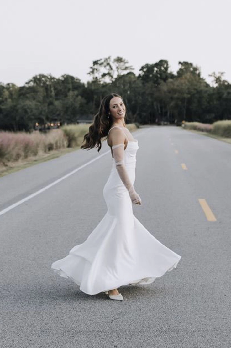 Bride twirls in a crepe fit and flare wedding gown with sheer sleeves, in the middle of an open road at dusk. 