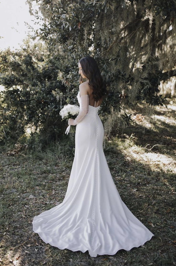 Back of bride in a crepe fitted gown with thin straps and sleeves, holding flowers in front of trees in the sunlight. 