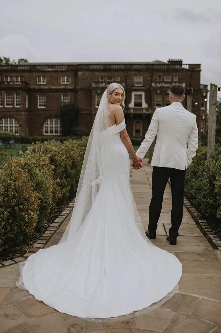 Bride looks past her shoulder in an off-shoulder draped gown with a veil, and holds hands with the groom outside the venue.