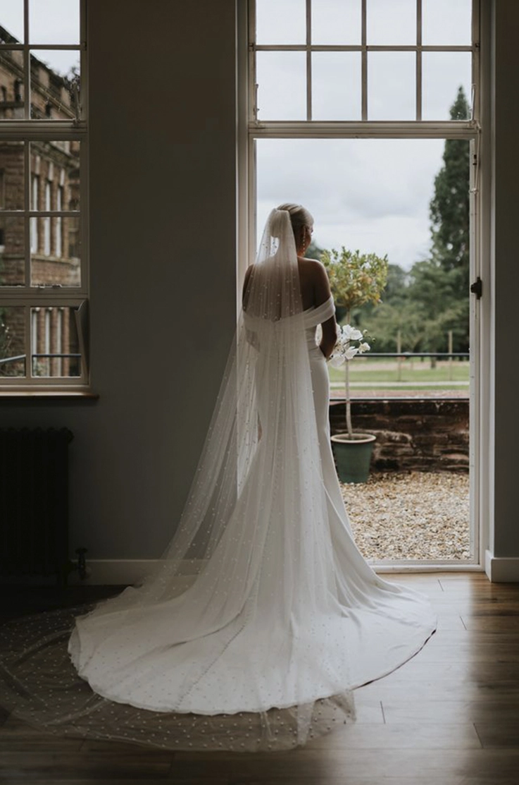Bride in an off shoulder crepe gown with a draped bodice and a veil, is holding flowers and gazes out French doors.