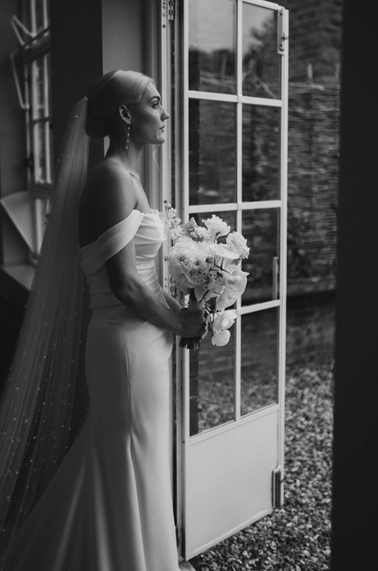 Bride stands in a doorway in a crepe fit and flare wedding dress with off-shoulder draped sleeves and a veil.