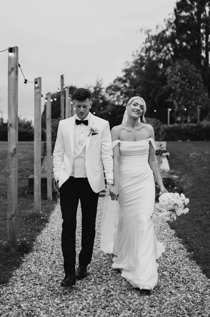 Bride in a scoop neck crepe gown with a draped bodice, walks hand in hand with her groom on an outdoor path.