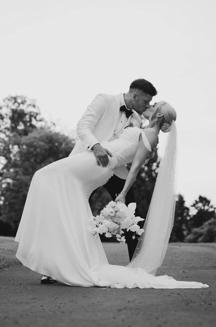 Groom dips his bride in a crepe fit and flare gown with draped sleeves and a veil, holding a white bouquet.
