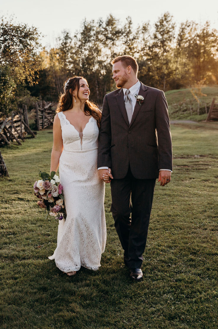 A bride in a sleeveless lace wedding dress holds a bouquet of flowers and walks with a groom in a suit while looking at each other, outside in a grass field with trees in the background.