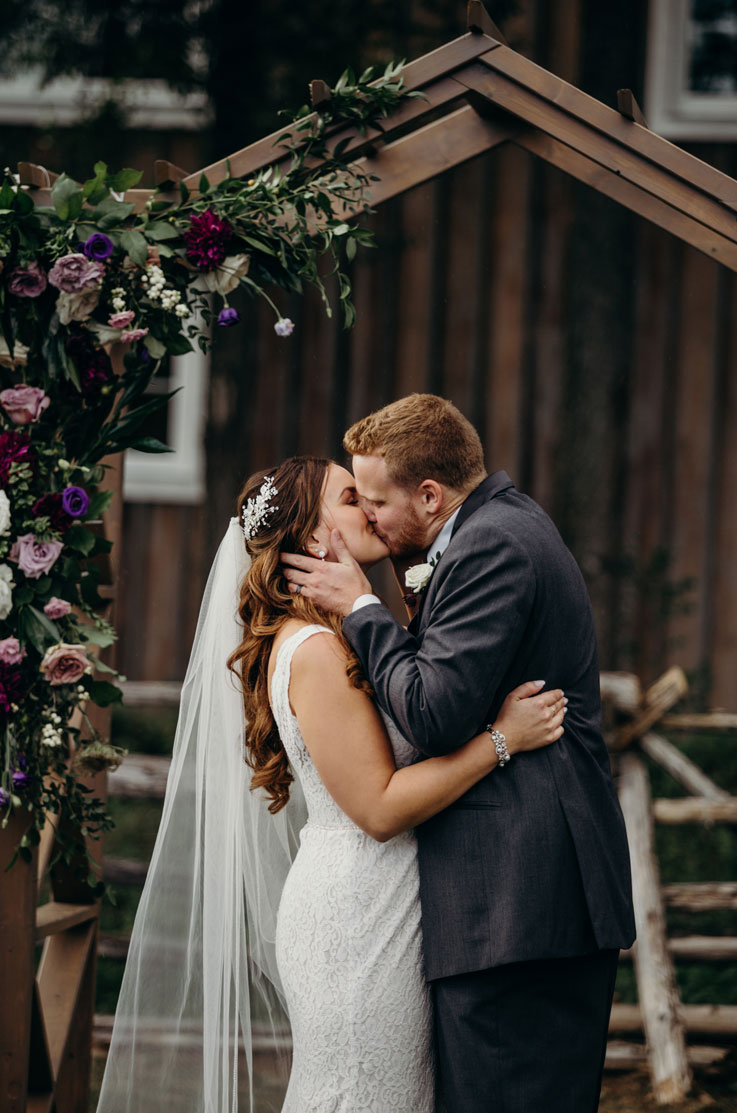 A bride in a sleeveless lace wedding dress and a veil, and a groom in a suit hold each other and kiss in front of a geometric wedding ceremony decoration outdoors.
