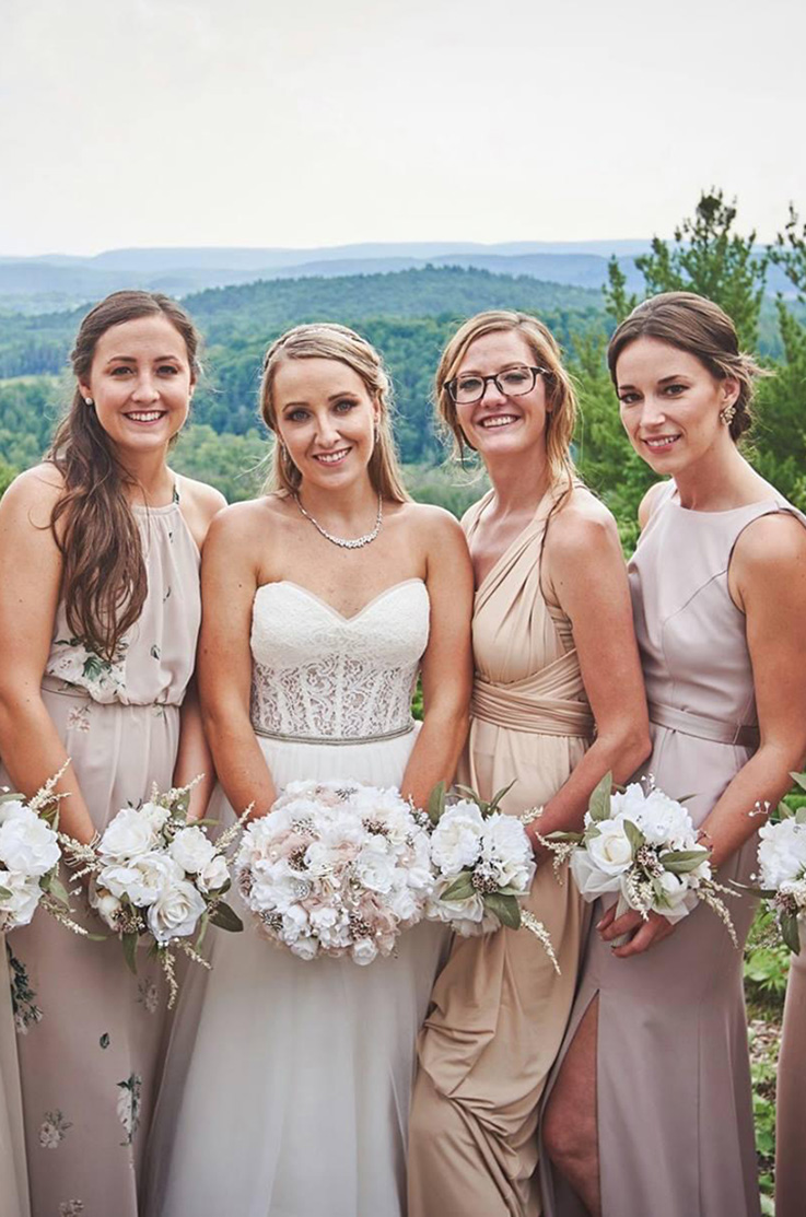 A bride in a sweetheart neckline wedding dress stands with three bridesmaids, all of them holding bouquets, outdoors with the view of rolling hills and forest behind them.