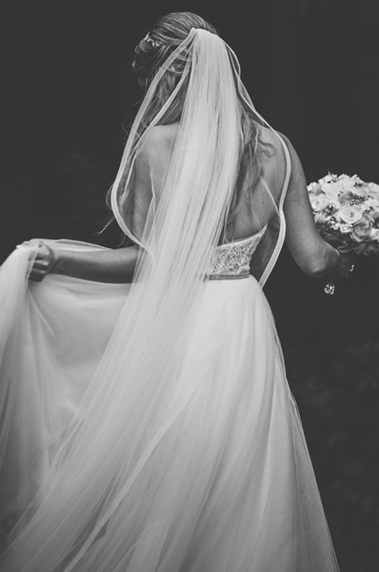 A bride in a lace and tulle wedding dress and a veil stands and faces away, with a bouquet in one hand while holding the side of her skirt up with the other.