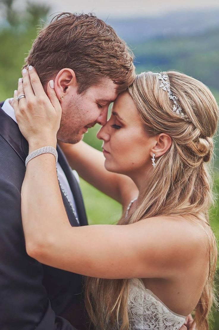 A bride in a lace wedding dress and a groom in a suit hold each other close with their eyes closed and foreheads touching, while standing outside.