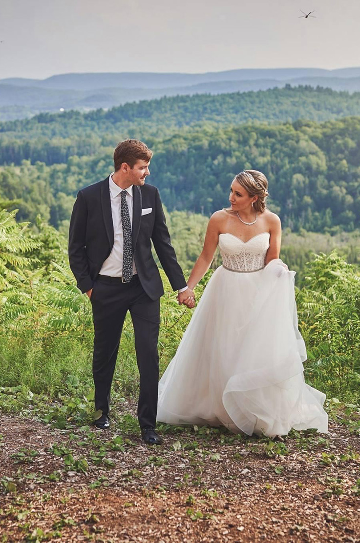 A bride in a sweetheart neckline wedding dress and a groom in a suit hold hands and look at each other, while standing outside, with the view of rolling hills and forest behind them.