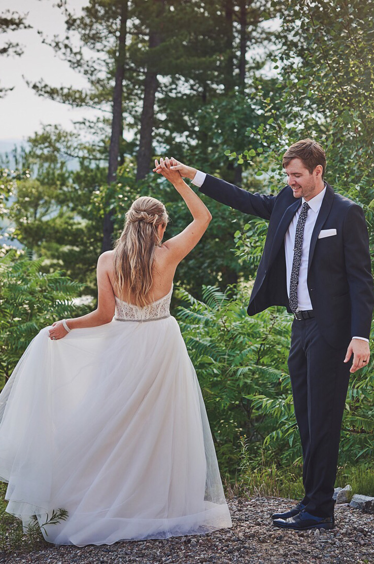 A bride in a lace and tulle wedding dress is being spun around by a groom in a suit outdoors in a forest.