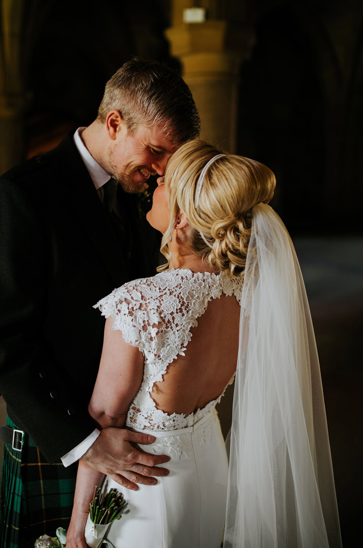A bride in a keyhole-back wedding dress and a veil, and a groom in a kilt look at each other, smiling, in a dim building.