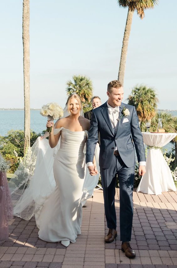 Bride in an off shoulder fit and flare gown with a veil, holds up a bouquet as she walks down the aisle with her groom. 