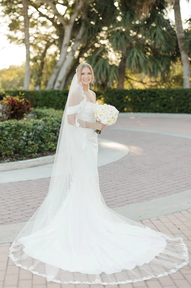 Bride smiles in a crepe fit and flare gown with a draped bodice and sleeves, holding a bouquet by palm trees. 