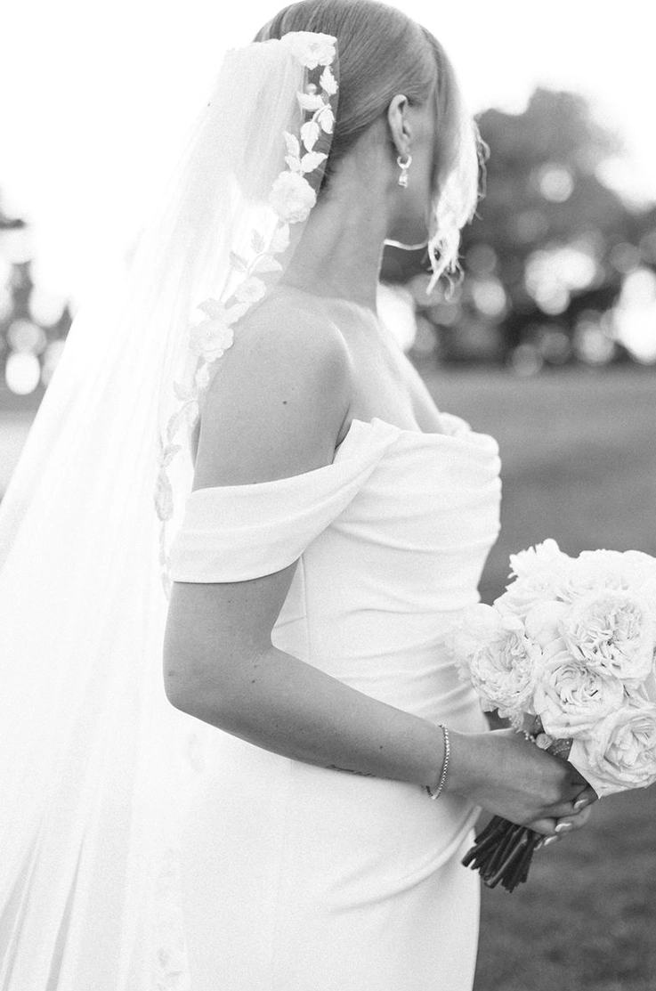 Close-up of bride in a draped crepe bodice gown with sleeves and a lace-trim veil, is holding a bouquet in black and white.