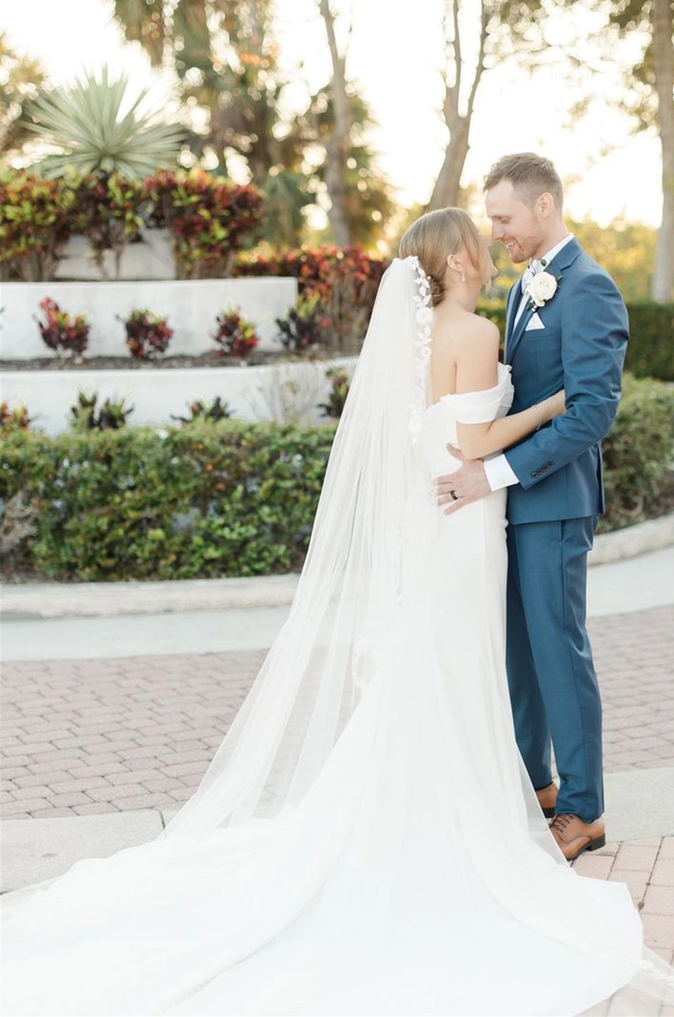 Groom holds his bride in an embrace wearing a draped crepe bodice gown with off-shoulder sleeves and a veil.