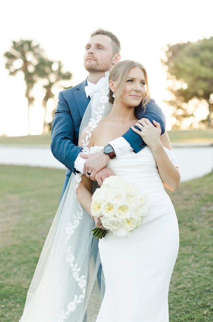 Groom in blue holds his bride in an embrace in a crepe fit and flare gown with a veil, holding a bouquet in front of palm trees. 
