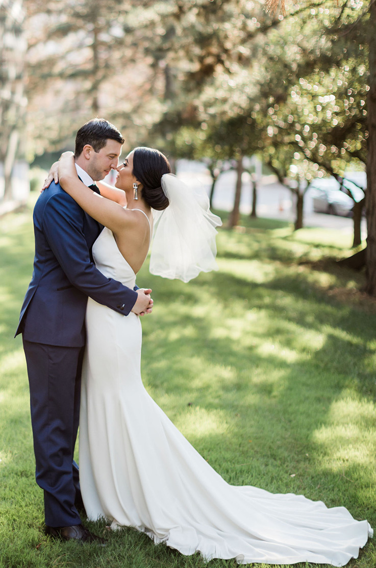 A bride in a fitted wedding dress and a veil, and a groom in a suit hold each other close and look into each others eyes, while standing outside with trees in the background.