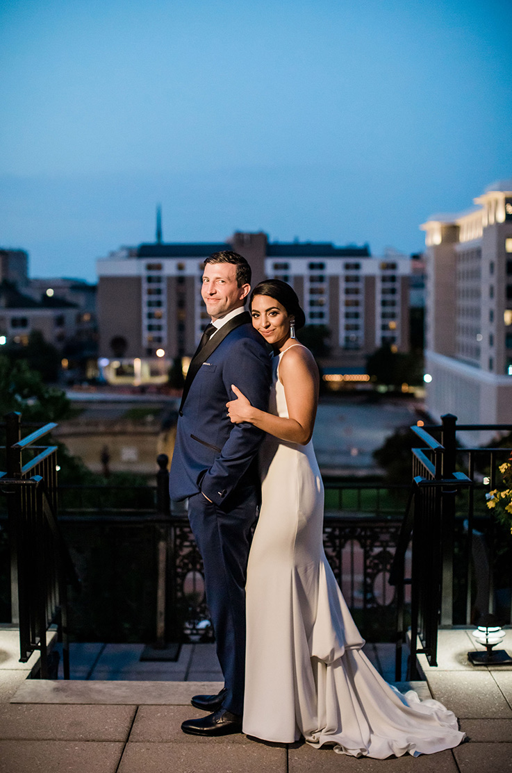 A bride in a fitted wedding dress stands behind a groom in a suit, her head on his shoulder, holding his arm, both of them smiling with the view of a city behind them.