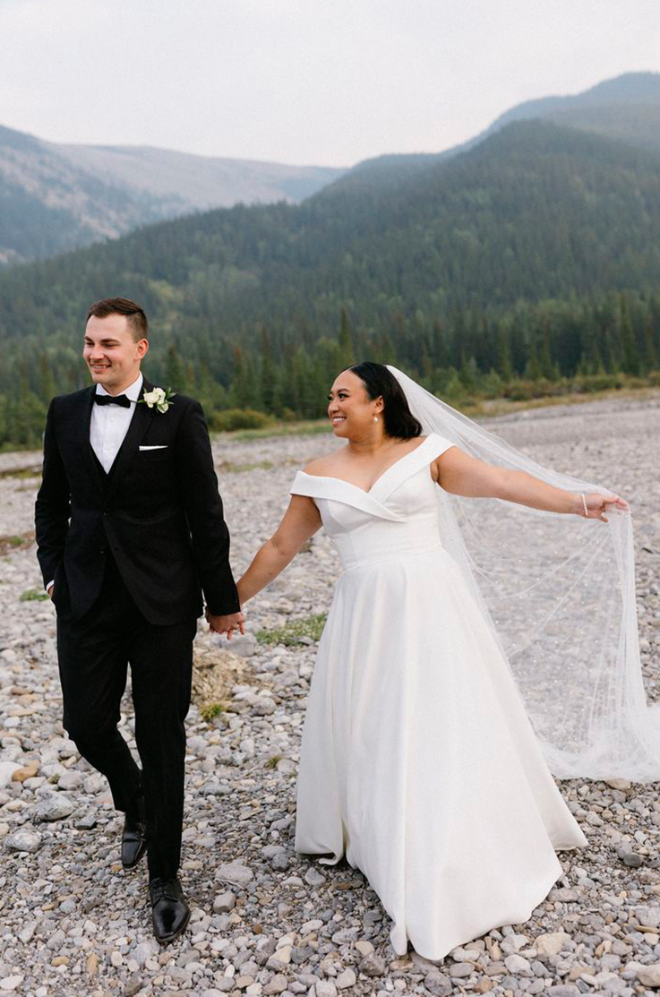 Bride in an off-shoulder A-line gown, holds up her veil and walks beside her groom in a tuxedo on rocks in front of mountains.
