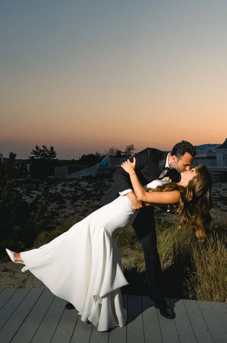 room dips his bride in a sleeveless crepe wedding dress with princess seams, standing on a boardwalk at sunset.
