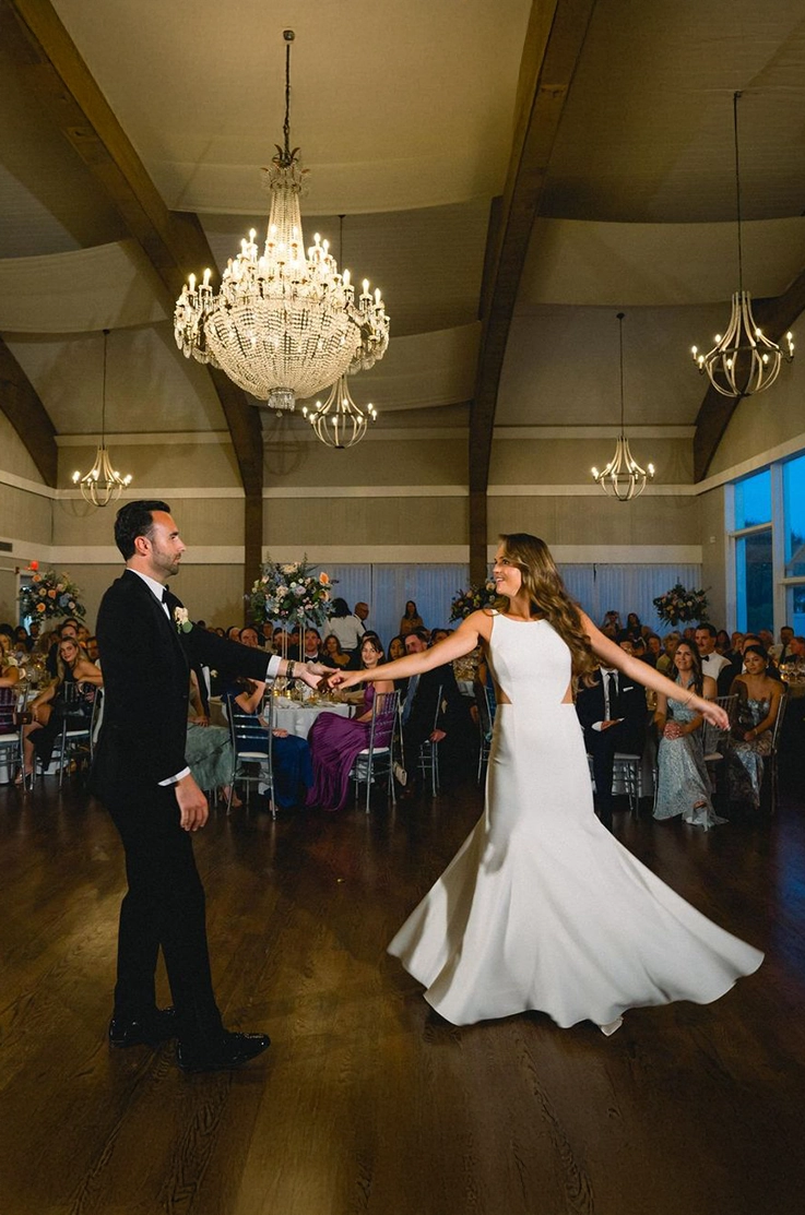 Groom dances with bride in a crepe fit and flare gown with a jewel neck, under chandeliers in front of guests sitting down.