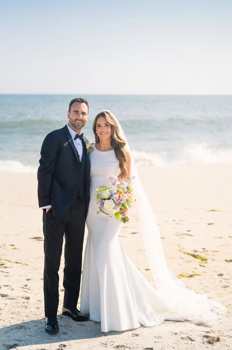 Bride in a fitted crepe sleeveless gown with a high neckline and a veil, is holding a bouquet beside groom on a beach.