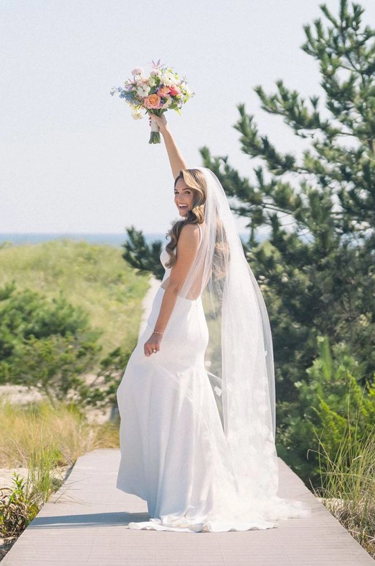Bride raises a colorful bouquet up, on a boardwalk in a crepe gown with a low open back and veil. 