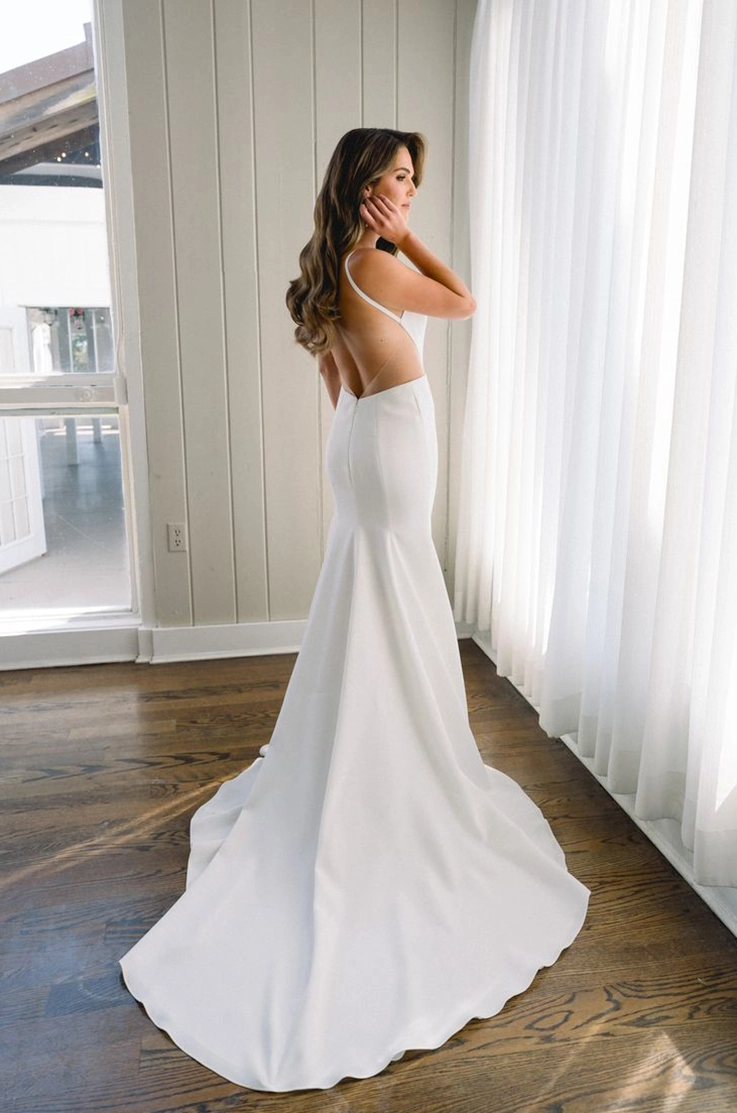 Back of bride posing in a sleek open-back crepe gown with thin straps indoors, by a bright window with a curtain. 