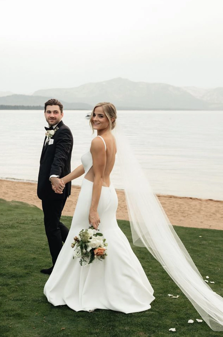 Bride holds hands with her groom and is walking in a sleek crepe wedding dress with a low open back and a veil outdoors.