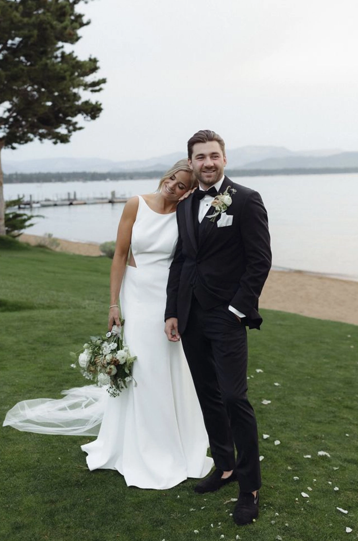 Groom kisses his bride in a fit and flare crepe gown with a low back and princess seams, standing near a lake.