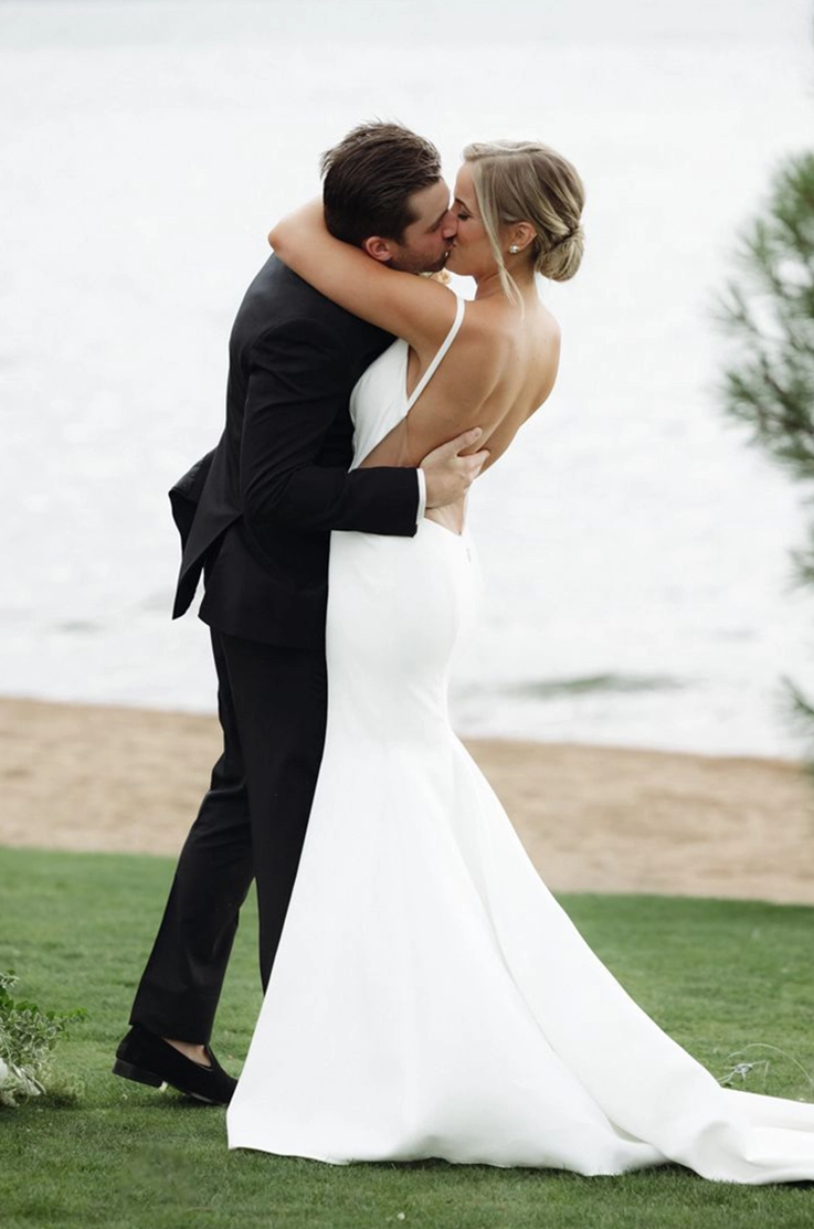 Groom kisses his bride in a fit and flare crepe gown with a low back and princess seams, standing near a lake.