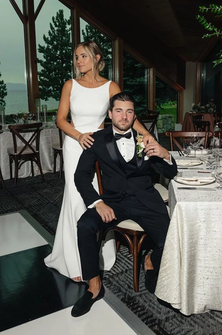 Bride in a crepe fit and flare gown poses behind groom sitting in a chair at the ceremony.
