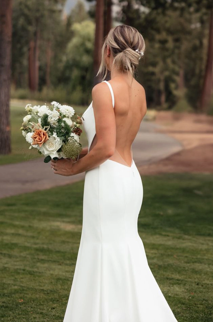 Back of bride in a fit and flare crepe gown with an open back and princess seams, poses with a bouquet. 