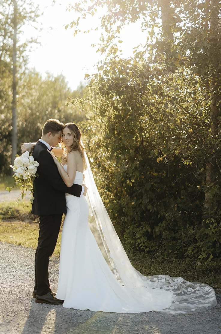 Bride in a charmeuse satin wedding gown with a veil embracing groom on a gravel path in the sunlight and holds a bouquet.