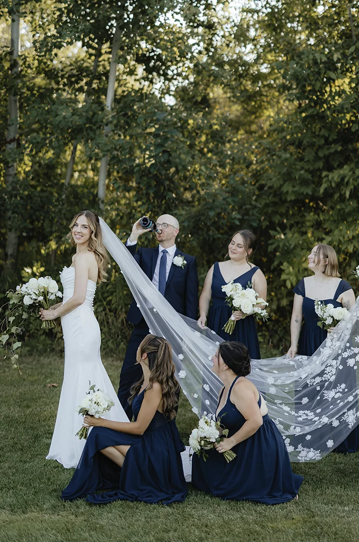 Bride in a charmeuse satin strapless gown with a draped bodice and a veil, being held by bridesmaids in navy dresses outdoors.  