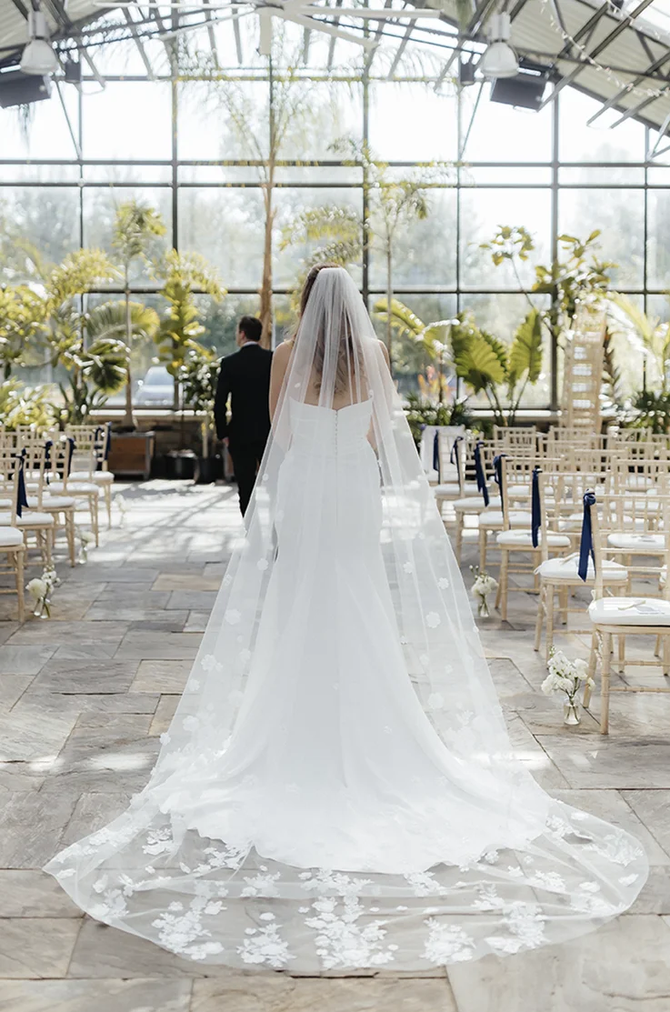 Bride in a strapless satin wedding dress with a floral lace veil walks towards groom, facing large windows in a greenhouse. 