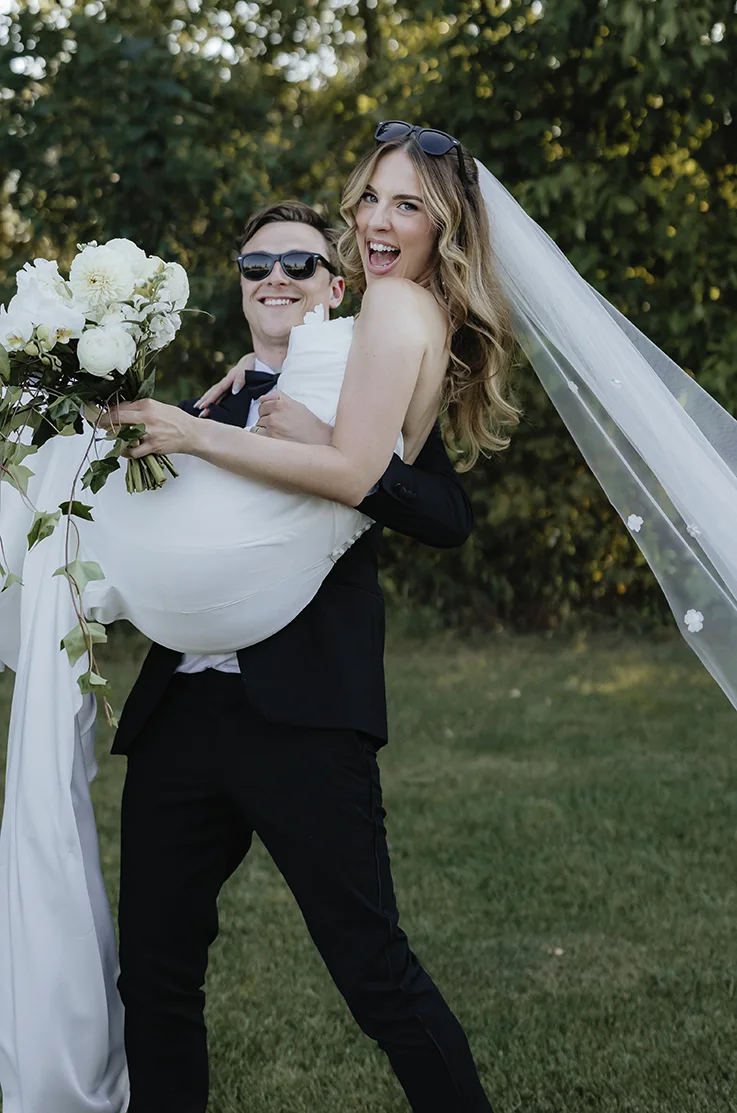Groom picks up his bride in a draped bodice gown with sequin lace detail and a veil, holding a white and green bouquet.