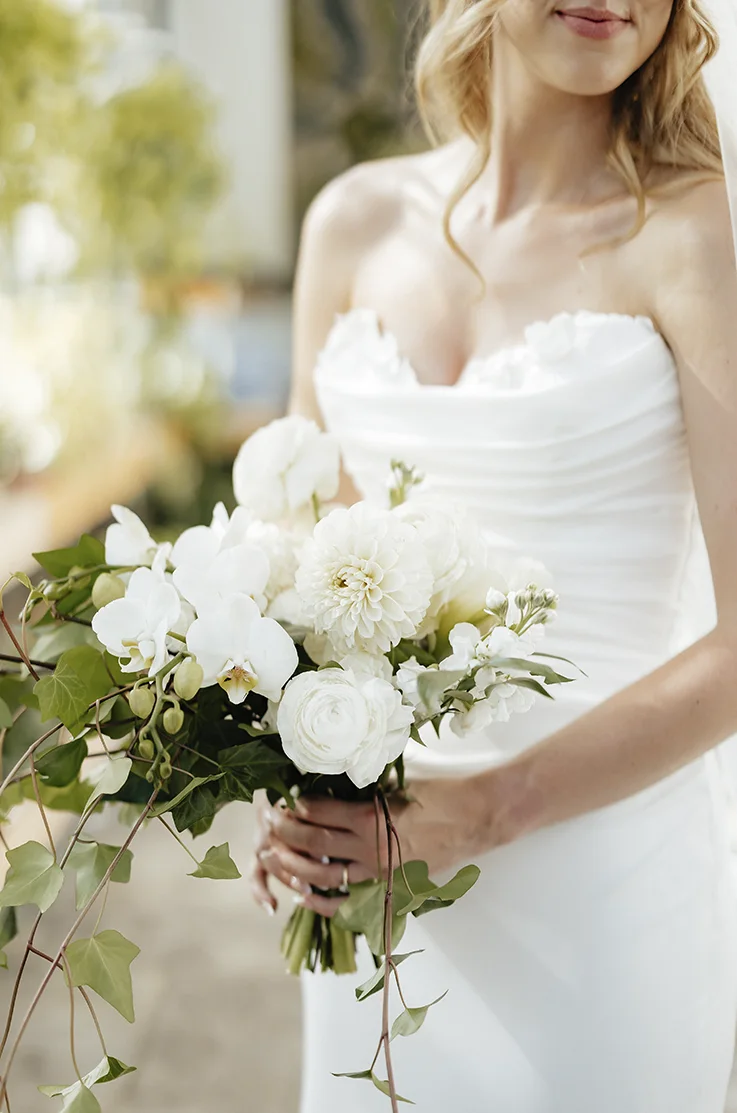 Close-up detail of bride in a strapless draped bodice gown with sequin lace and a veil, is holding a bouquet outdoors.