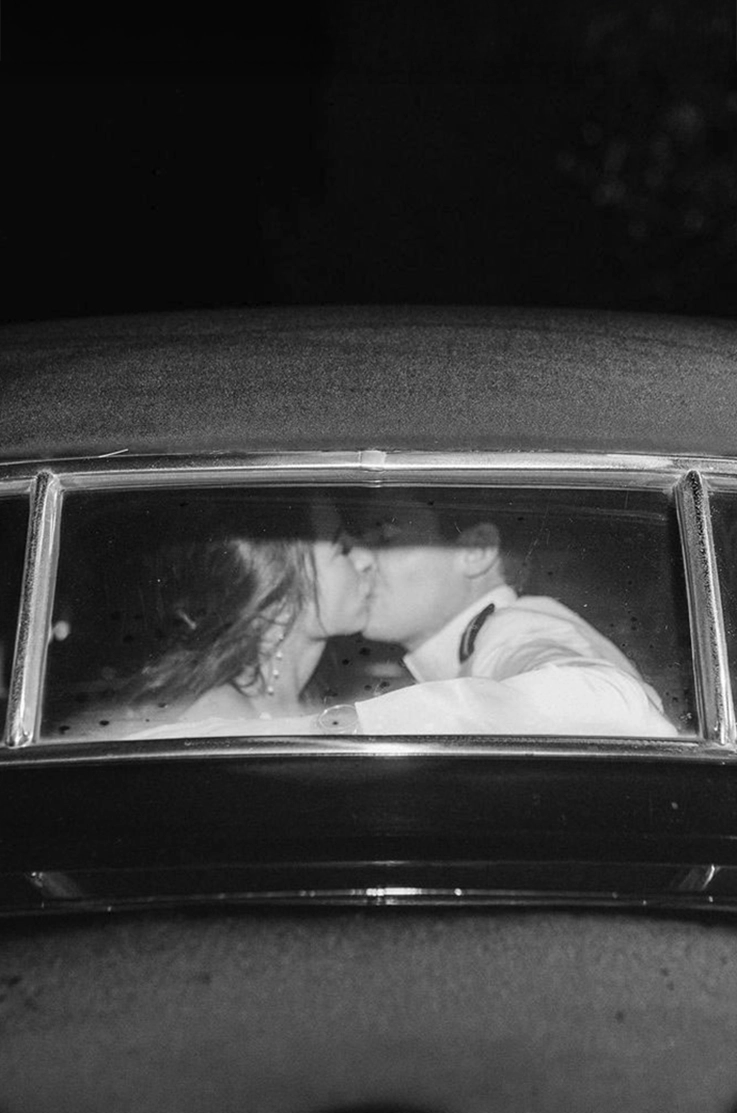 Newlyweds kiss in the back mirror of a black vintage car, shown in black and white during their wedding celebration.