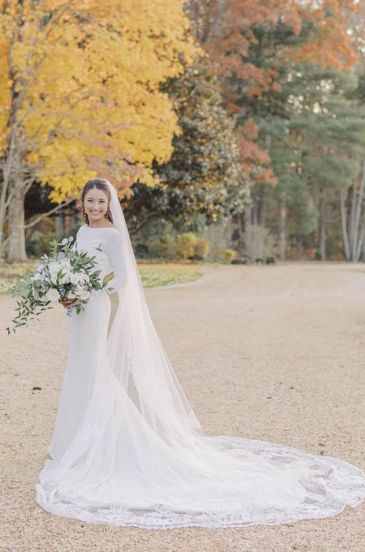 Bride in a long-sleeve crepe wedding dress with a bateau neck and a long veil, is holding a bouquet near autumn trees.