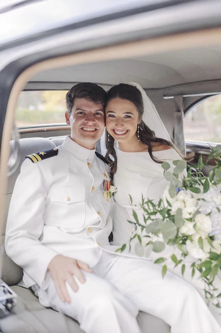 Bride in a bateau neck crepe gown with a veil, smiles next to groom in a car in uniform, and is holding flowers.