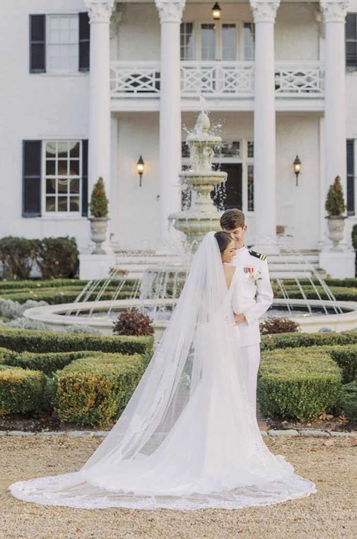 Bride in a long sleeve fitted gown embraces groom and poses in front of a grand fountain.